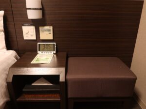 ドーミーイン鹿児島 ダブルルーム ベッドサイドテーブルと椅子 照明とエアコンのスイッチとACコンセント、目覚まし時計、ティッシュがあります