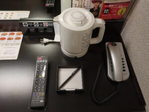 アパホテル＆リゾート 大阪梅田駅タワー スタンダードルーム デスク上 ACコンセント、有線LANポート、明かりのスイッチ、テレビのリモコン、電気ポット、メモ帳、電話機があります