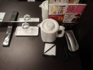 アパホテル 京都駅堀川通 シングルルームB デスク上 テレビのリモコン、電気ケトル、メモ帳、電話機があります