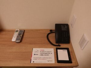 ドーミーイン水戸 ダブルルーム デスク上 ACコンセントと照明のスイッチ、電話機、メモ帳、テレビとエアコンのスイッチ、有線LANコネクタ